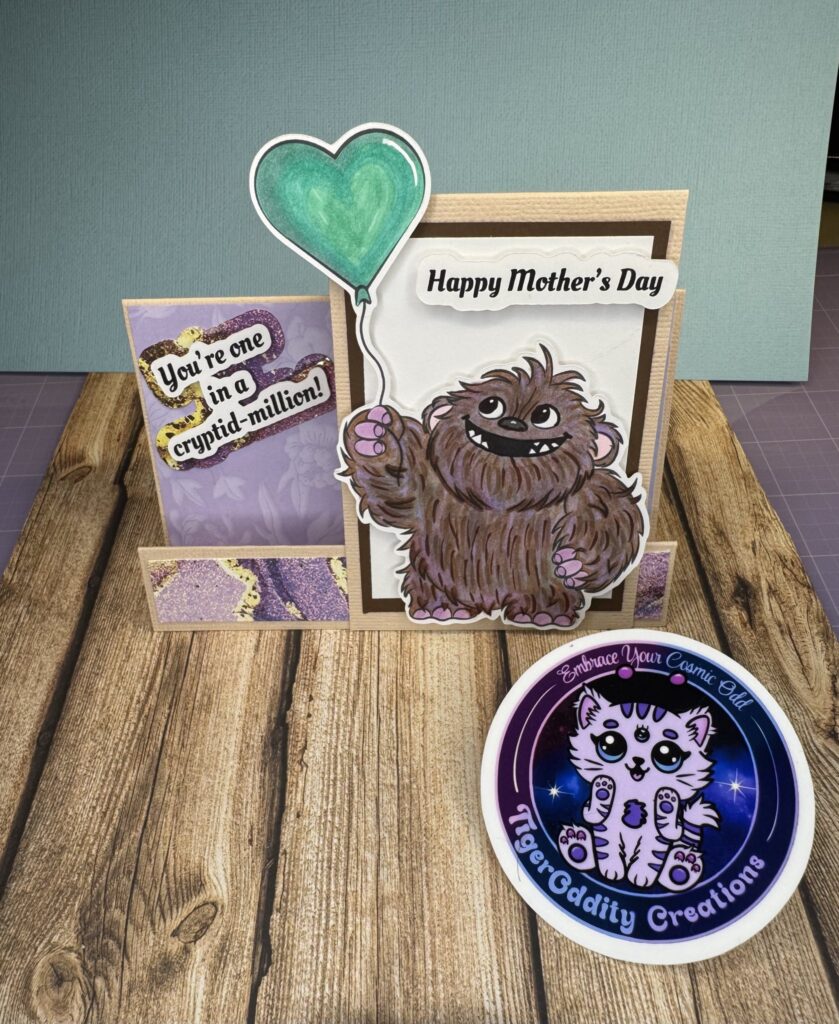 Handmade Mother’s Day card stair step fold featuring a cute Bigfoot/Sasquatch digital stamp named Woolly, holding a teal heart balloon. The card sentiment reads “You’re one in a cryptid-million!” Stamp is colored with Copic alcohol markers in brown and lavendar tones and uses a dimensional folded base for a 3D effect. Designed using a Freakin’ Cute Stamps image/digital stamp.